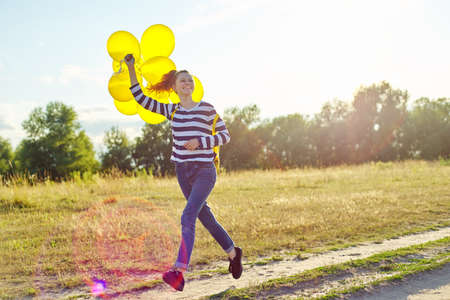 Happy teenager girl with yellow balloons and backpack running and jumping along country road in summer meadow. Freedom, life, joy, holiday conceptの写真素材