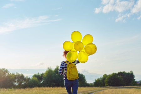 Happy teenager girl with yellow balloons and backpack running and jumping along country road in...