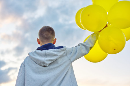 Teen boy running with balloons, view back. Nature, sky in clouds background. Holiday, birthday,...