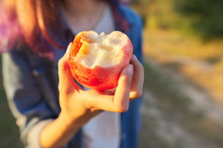 Tasty juicy eaten red apple in girls hand closeup, nature sunset golden hour backgroundの写真素材