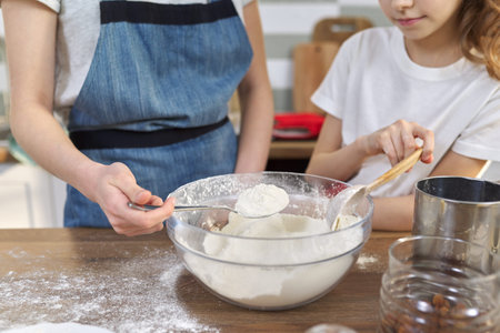 Two girls, teenager and younger sister, preparing cookies together in kitchen. Children mix flour in...