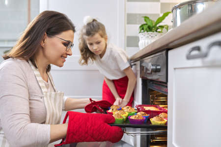 Mother in apron and daughter little helper preparing cupcakes together. Woman taking out tray with baked cupcakes from oven, interior home kitchen. Family, mothers day, healthy homemade baking foodの写真素材