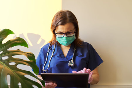 Portrait of doctor woman in medical protective mask with digital tablet in office. Light wall...