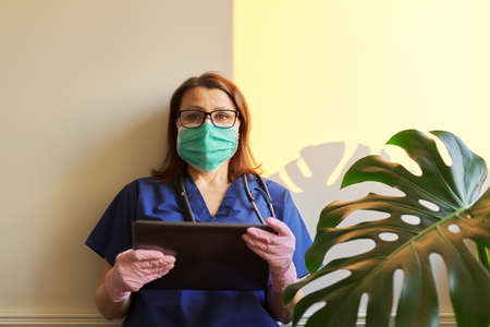 Serious woman doctor in medical blue uniform, protective mask, gloves reading digital tablet, medic standing in office near window. Medicine, healthcare, people conceptの写真素材