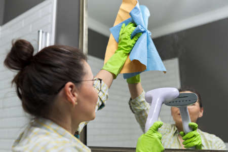Woman doing cleaning in bathroom using vacuum cleaner, without use of household chemicals. Close-up of gloved hands with steam in the mirror, eco-friendly cleaningの写真素材