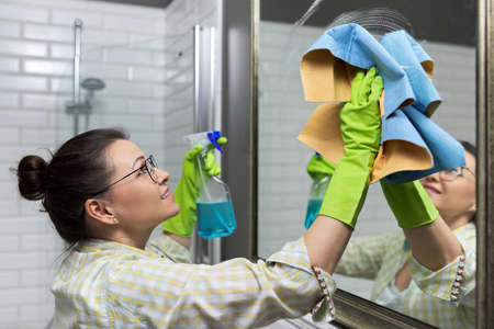 Woman cleaning mirror in bathroom using professional rag and washing spray, close upの写真素材