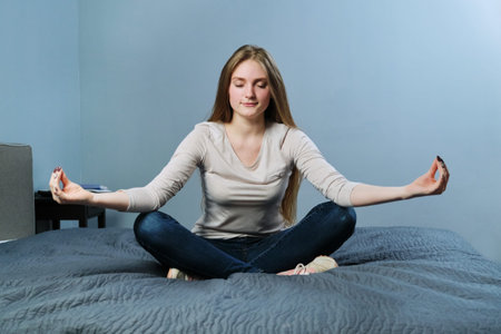 Relaxed young woman sitting at home in bed in lotus position, meditating girl practicing yoga