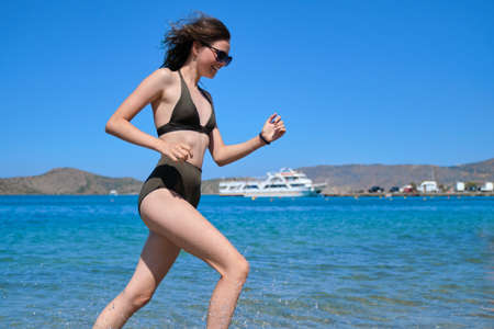 Beautiful laughing teenager girl in sunglasses swimsuit bikini running on sea water, sunny summer day. Relax, fun, vacation on the beachの写真素材