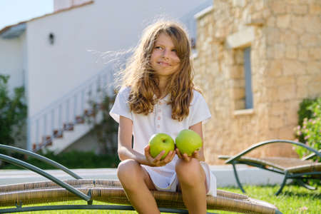 Outdoor portrait of a child girl with two green apples sitting in a garden chairの写真素材