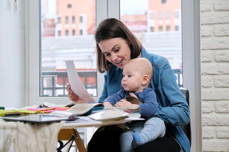 Young mother work with baby in her arms at home office. Woman interior designer working with fabric samples, on the table tablet computer, sketches and textile palletsの写真素材