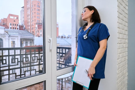 Female nurse with medical record standing in office looking out the window, female general...