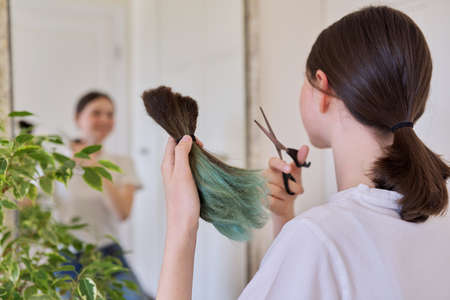 Girl makes a haircut at home, teenager cut her hair, in hands bunch of cut hair and scissors, cut dyed unhealthy hairの写真素材