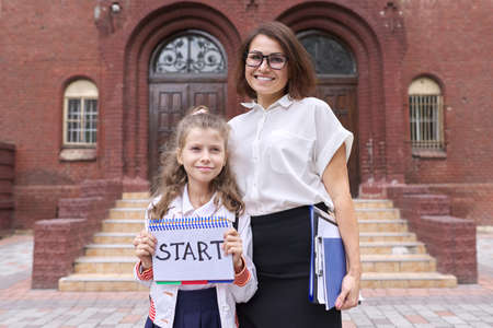 Start school, female teacher and girl student together, school building background. Girl holding notebook with handwritten word start. Back to school, education conceptの写真素材