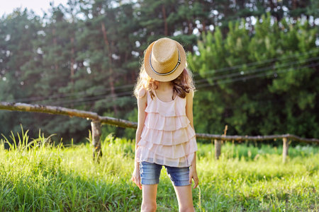 Happy girl enjoying summer vacation, kid in nature on green meadow playing with hat, covered her...