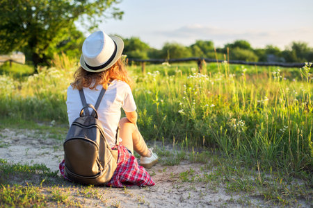 Happy teenager girl in hat with backpack sitting on country road, enjoying summer vacation,...