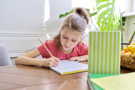 Girl child studying at home, sitting at table. Schoolgirl writes in notebook, looks into book,...