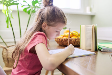 Kid girl studying at home, sitting at the desk, writing in school notebook. Homework, distance...