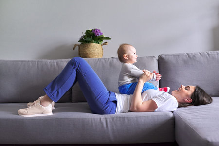 Happy mother with toddler son at home. Laughing woman playing with baby lying on sofa. Family,...