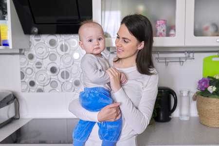 Happy mother with toddler son at home. Smiling woman holding 1 year old baby in her arms in kitchen....