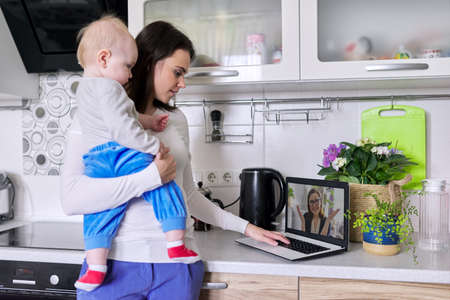 Young mother with toddler in her arms talking online with psychologist, making video call, video conference using laptop at home. Technology, mental health, therapy, distance lifestyle conceptの写真素材