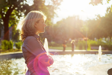 Portrait of a beautiful mature woman in profile, copy space, city fountain in sunset sunlight backgroundの写真素材