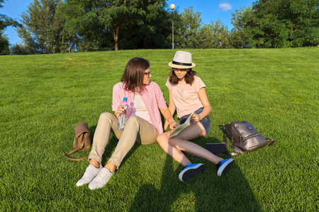 Mom and daughter teenager student together sitting on the green lawn having a rest, girl showing...