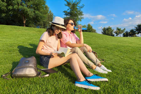 Mom and daughter teenager student together sitting on the green lawn having a rest, girl showing mother a book, talking parent and childの写真素材