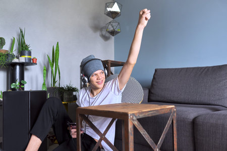 Young trendy hipster in headphones listening to music, teen boy with smartphone sitting at home...