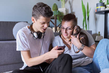 Happy talking mom and teenage son, parent and child sitting at home on the floor with pet little dog toy terrier, real friendly loving familyの写真素材