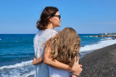 Happy mom and daughter kid hugging walking together along the beach, back view. Family, vacation, happiness, childhood, mother and child communicationの写真素材