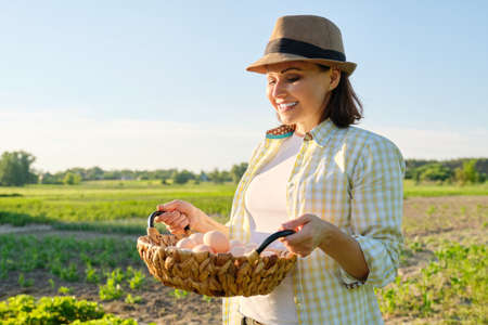 Portrait of farmer woman in hat with basket of fresh egg. Agriculture, farming organic eco products. Country road, countryside, sunset, nature, garden backgroundの写真素材
