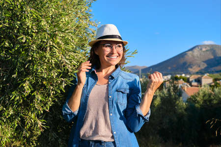 Portrait of mature beautiful smiling woman in hat, jeans clothes, copy space. Nature, mountains, olive trees, sky, sunset landscape backgroundの写真素材
