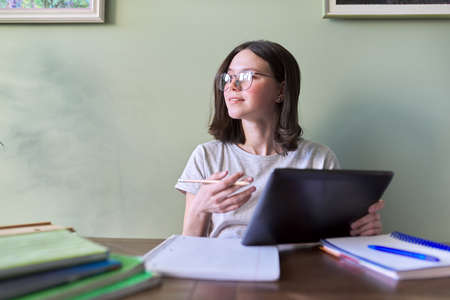 Teenage girl studies at home, sits at table with books and notebooks using digital tablet. Technologies in education, online learningの写真素材