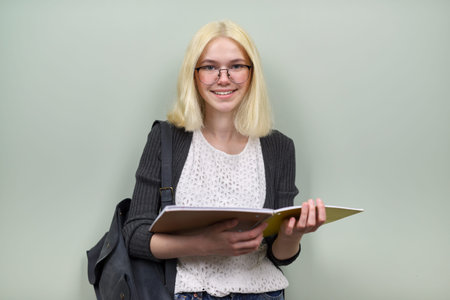 Portrait of happy smiling student girl teenager 16 years old in glasses with backpack textbooks looking at camera, on green color backgroundの写真素材