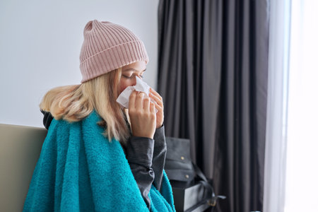 Teenager girl sneezing in handkerchief, female with symptoms of illness sitting at home covered with warm blanket and wearing knitted hatの写真素材