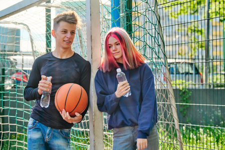 Active healthy lifestyle of teenagers 16, 17 years old in city. Couple with trending hairstyles on sports basketball court talking, standing with ball and bottle of waterの写真素材