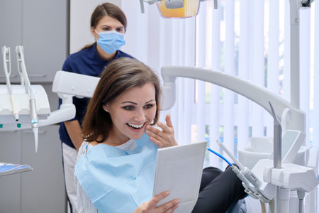 Happy middle aged woman with doctor dentist looking in mirror at teeth, sitting in dental chair. Medicine, dentistry and health care conceptの写真素材