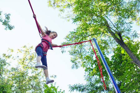 Outdoor active recreation and amusement park, child girl having fun jumping on trampoline with elastic ropesの写真素材