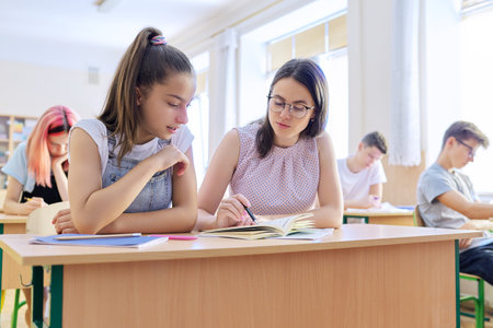 Young woman teacher teaches lesson in classroom of teenage children, teacher sits at desk with student, checks knowledge. Education, school, college, teaching conceptの写真素材