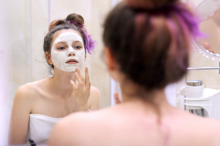 Young beautiful woman with white cosmetic natural healing mask on face in bathroom. Cosmetology, moisturizing and facial in adolescence, clean and perfect skinの写真素材