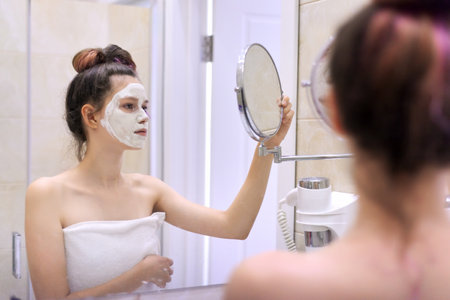 Young beautiful teen girl with white cosmetic natural healing mask on face in bathroom. Cosmetology, moisturizing and facial in adolescence, clean and perfect skinの写真素材