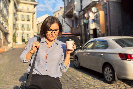 Mature business woman in glasses with office laptop bag walking along city street. Happy smiling middle aged female with cup of coffee, urban style backgroundの写真素材