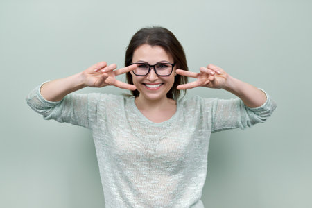 Happy mature confident woman showing victory symbol with hand gesture smiling looking at camera on green background. Middle aged woman housewifeの写真素材