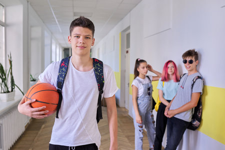 Portrait of student teenager guy 16, 17 years old with basketball ball backpack in school building, group of students talking in corridor background. Teens, education, sport, active healthy lifestyleの写真素材