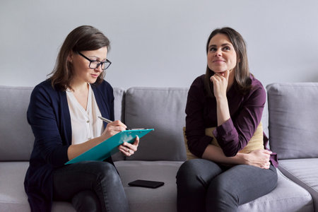 Social worker, psychologist talking to young woman in office. Women sitting on couch, counselor making notes, listening to patient. Psychology, mental health, social difficulties of young peopleの写真素材