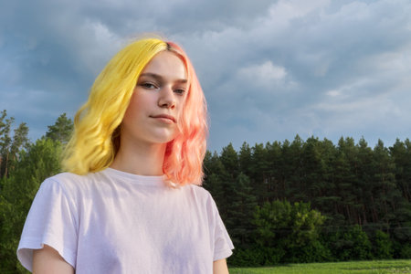 Portrait of serious teenage girl looking at camera, nature dramatic sky in clouds background. Teenager with trendy dyed colored hairstyleの写真素材