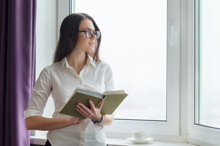 Young smiling woman in glasses reading a book, female near window with cup of coffee. Knowledge, rest, education, lifestyleの写真素材