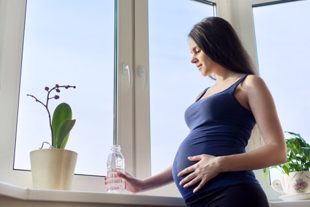 Young beautiful pregnant woman drinking water from bottle standing at home near the window, in third trimester of pregnancy, active healthy lifestyle, copy spaceの写真素材