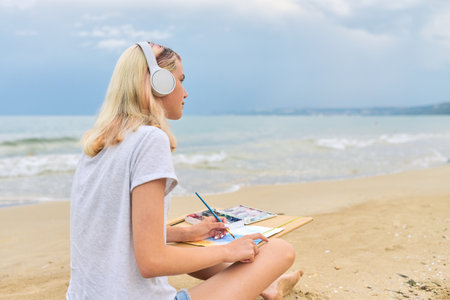 Young woman artist painting with watercolors sitting on the seashore, creative female in headphones listening to music, beautiful seascape background, copy spaceの写真素材