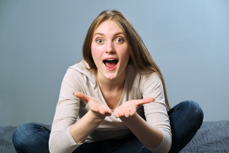 Happy beautiful young blonde woman with long hair smiling emotionally with open mouth, holding hands in front of face, copy space. Female sitting in lotus position. Emotion love, happiness, joyの写真素材